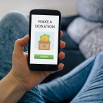Closeup Of A Person’s Hand Holding A Phone, Promoting Charitable Donations For Tax Deductions.