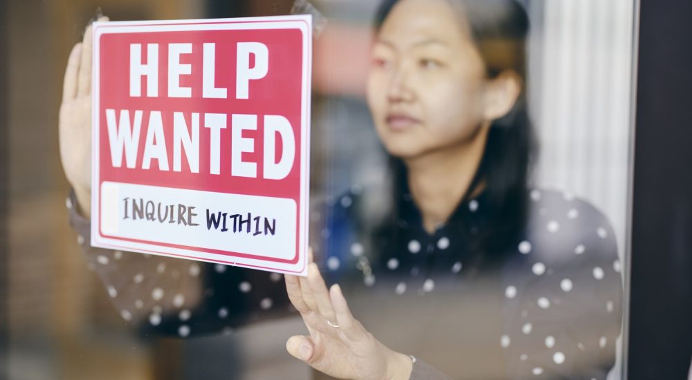 A Small Business Owner Putting Up A Help Wanted Sign In Her Store Window