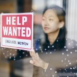 A Small Business Owner Putting Up A Help Wanted Sign In Her Store Window