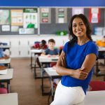 A Teacher In A Classroom Leans On A Desk And Smiles With Students In The Background