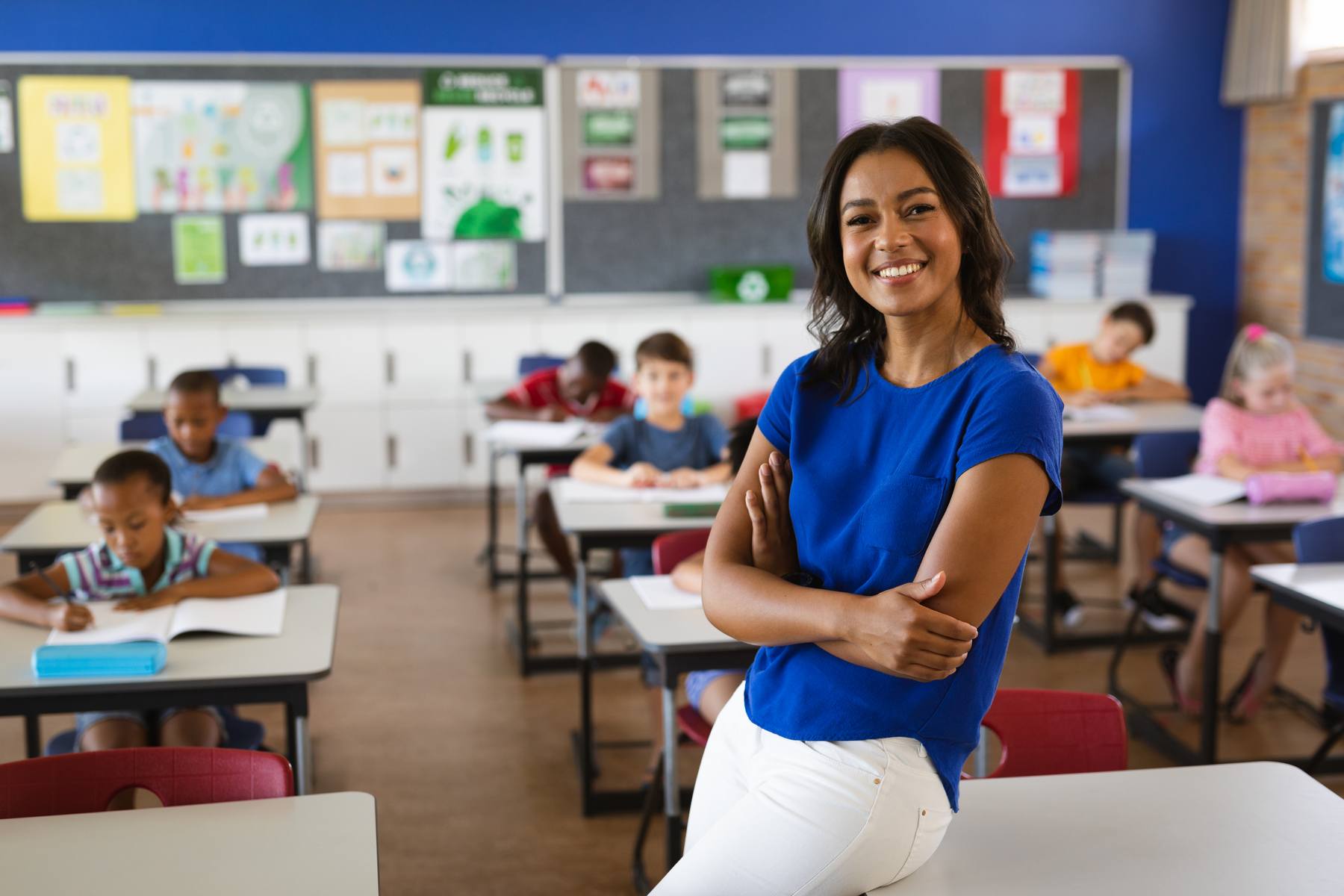 A teacher in a classroom leans on a desk and smiles with students in the background