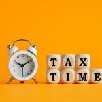 A White Analog Alarm Clock Rests Next To Alphabet Blocks Reading “tax Time” Before An Orange Background