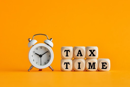 A white analog alarm clock rests next to alphabet blocks reading “tax time” before an orange background