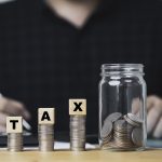 Three Stacks Of Coins In Increasing Height With Wood Blocks Spelling TAX On Top, Leading To A Coin Jar