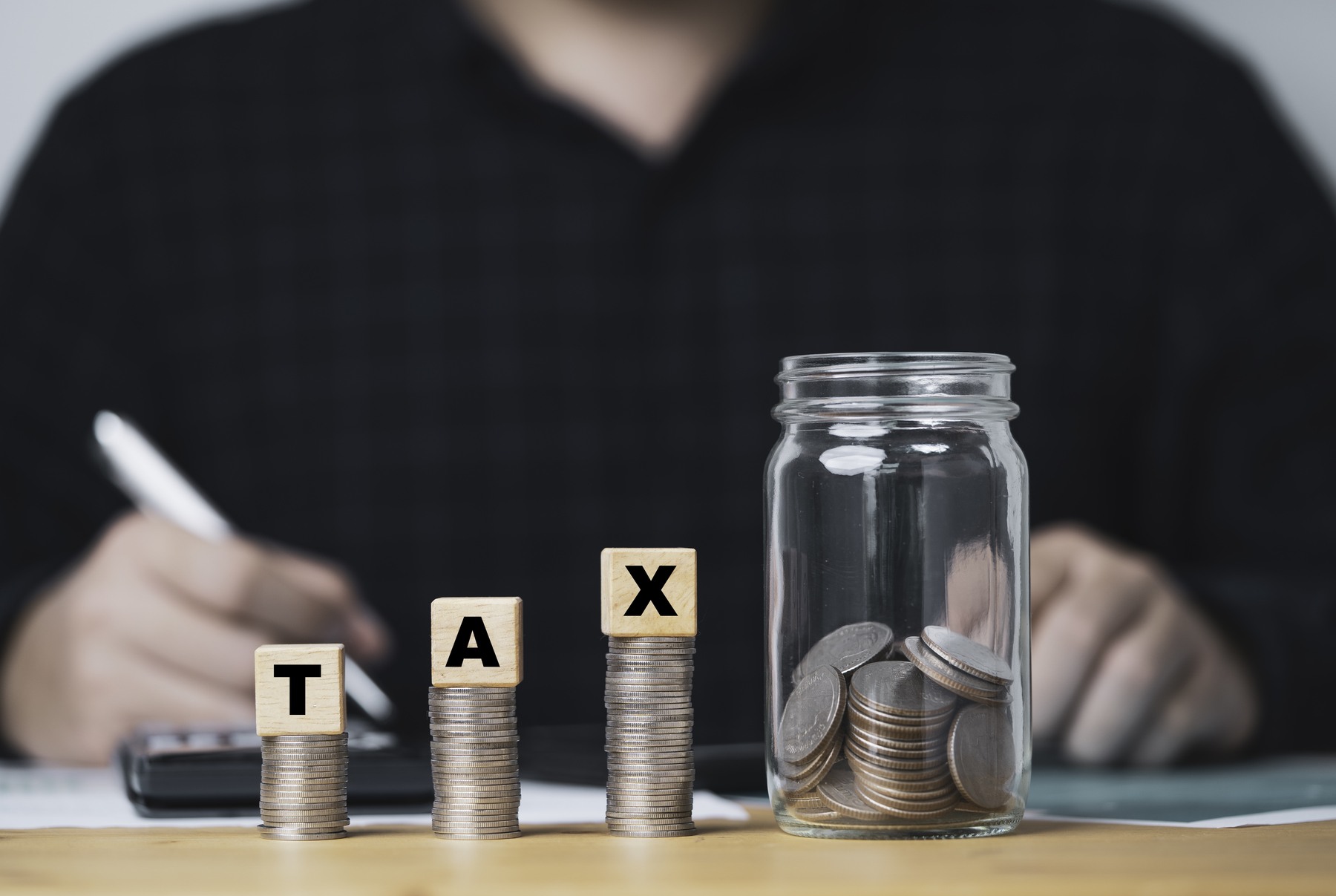 Three stacks of coins in increasing height with wood blocks spelling TAX on top, leading to a coin jar