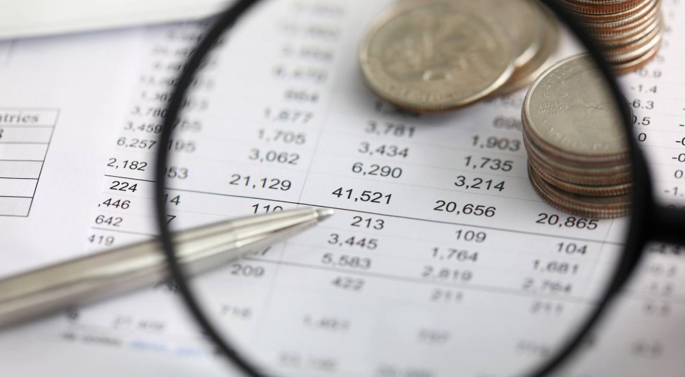 View Of Financial Fraud Ledger Details In A Table Through A Magnifying Glass With Coins In The Background