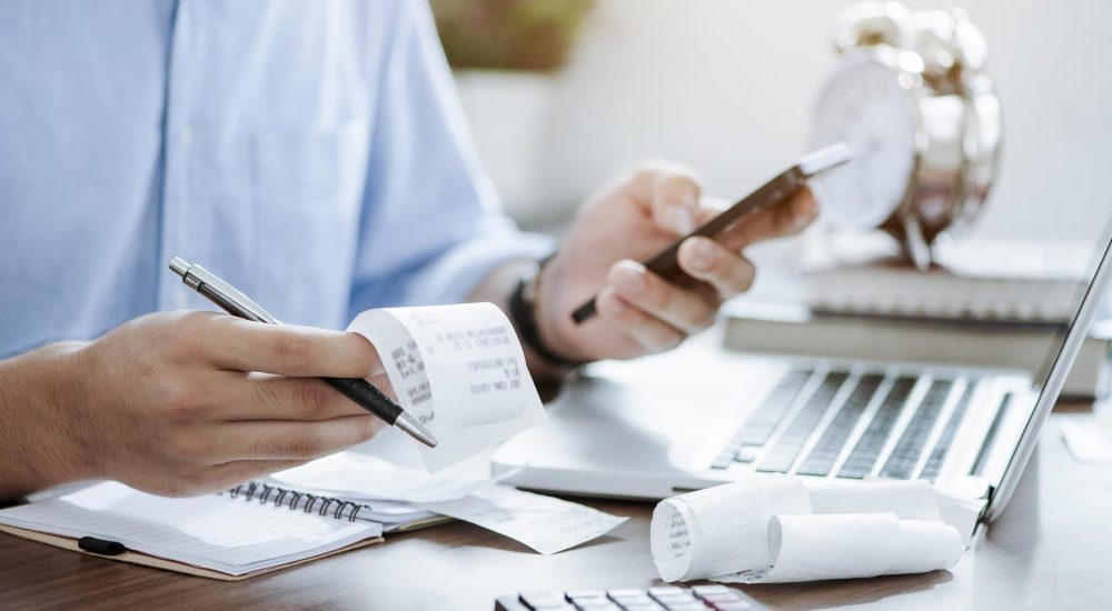 Man Holding Pen With Bills, Receipts, Laptop, And Calculator On His Desk